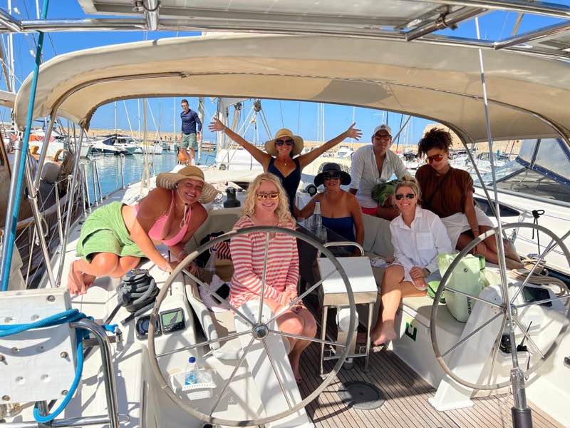 Smiling travelers seated in the cockpit of a sailboat in a marina, one with arms raised