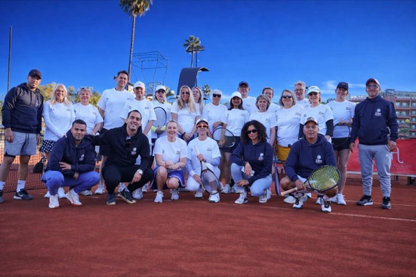 Travelers in white Elite shirts and navy hoodies pose with rackets on a red clay court under palm trees and a blue sky